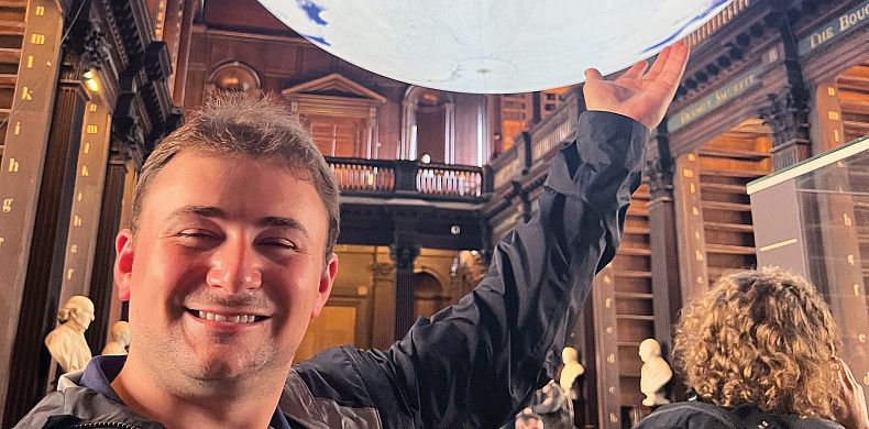 Person smiling indoors, seemingly holding up a large globe installation in a library setting. Books and busts are visible in the background.