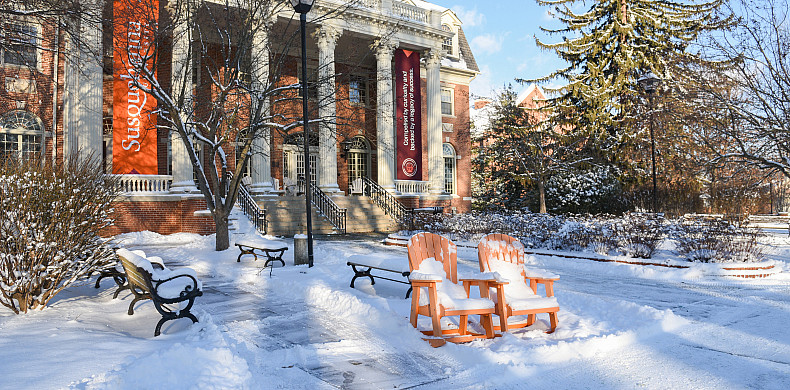 Image of two orange adirondack chairs covered in snow with a brick and column building in the background. Snow is on the ground.