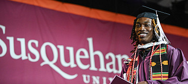 A graduate in a maroon gown and cap stands smiling, holding a diploma. Behind them is a banner with Susquehanna University.